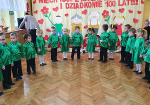 W pólkolu dziewczynki w zielonych sukienkach oraz chłopcy w zielonych koszulach i czarnych spodniach śpiewają piosenkę dla babci i dziadka